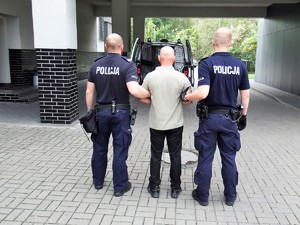 dwóch policjantów przytrzymuje zatrzymanego mężczyzna, który ma założone kajdany zespolone