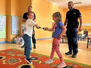 dwójka dzieci ma założone kajdanki, uśmiechają się, a za nimi stoją dwaj policjanci, którzy prowadzą spotkanie profilaktyczne