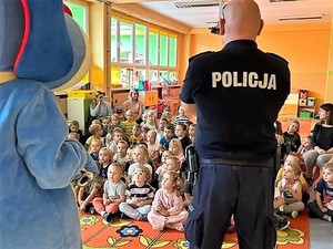 policjant stoi przodem do dzieci, prowadzi spotkanie profilaktyczne, obok niego stoi maskotka Śląskiej Policji