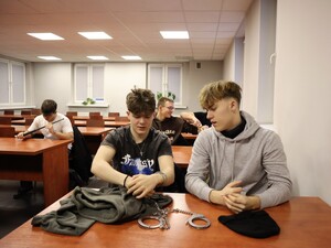 praktykanci siedzący w sali odpraw oglądają kajdanki i pałki służbowe
