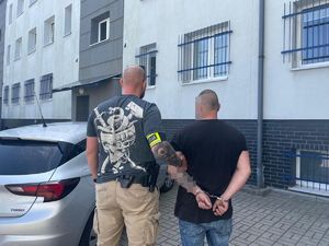 policjant trzyma zatrzymanego mężczyznę, stoją tyłem, zatrzymany ma na rękach założone kajdanki