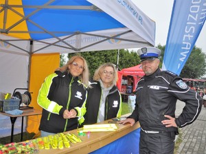 policjant pozuje do zdjęcia z pracownicami Starostwa Powiatowego w Wodzisławiu Śląskim, stoją przy stoisku profilaktycznym, na stole leżą odblaskowe gadżety