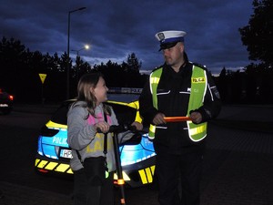 policjant rozmawia z dziewczyną, która trzyma hulajnogę, ma założone odblaskowe opaski na nadgarstki