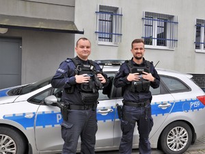 do zdjęcia pozują dwaj policjanci, stoją przed radiowozem